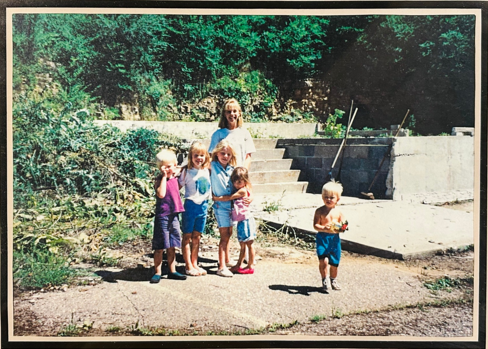 A 1990s family photograph of children standing on the bare cement slab among the trees, block walls and steps behind them