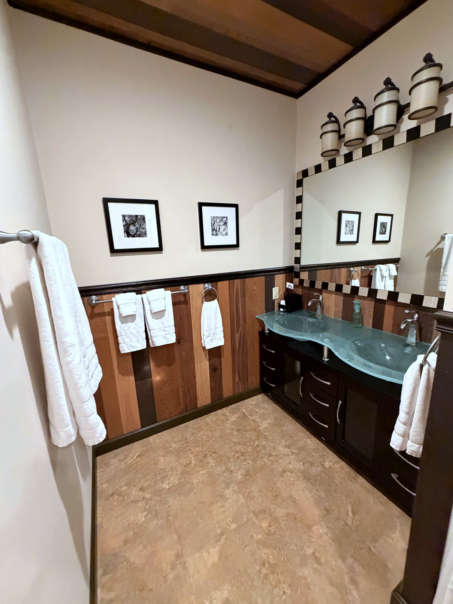 The third bath, seen wider: the double glass sinks under the checkered mirror, framed prints and towels on the wood-plank wall