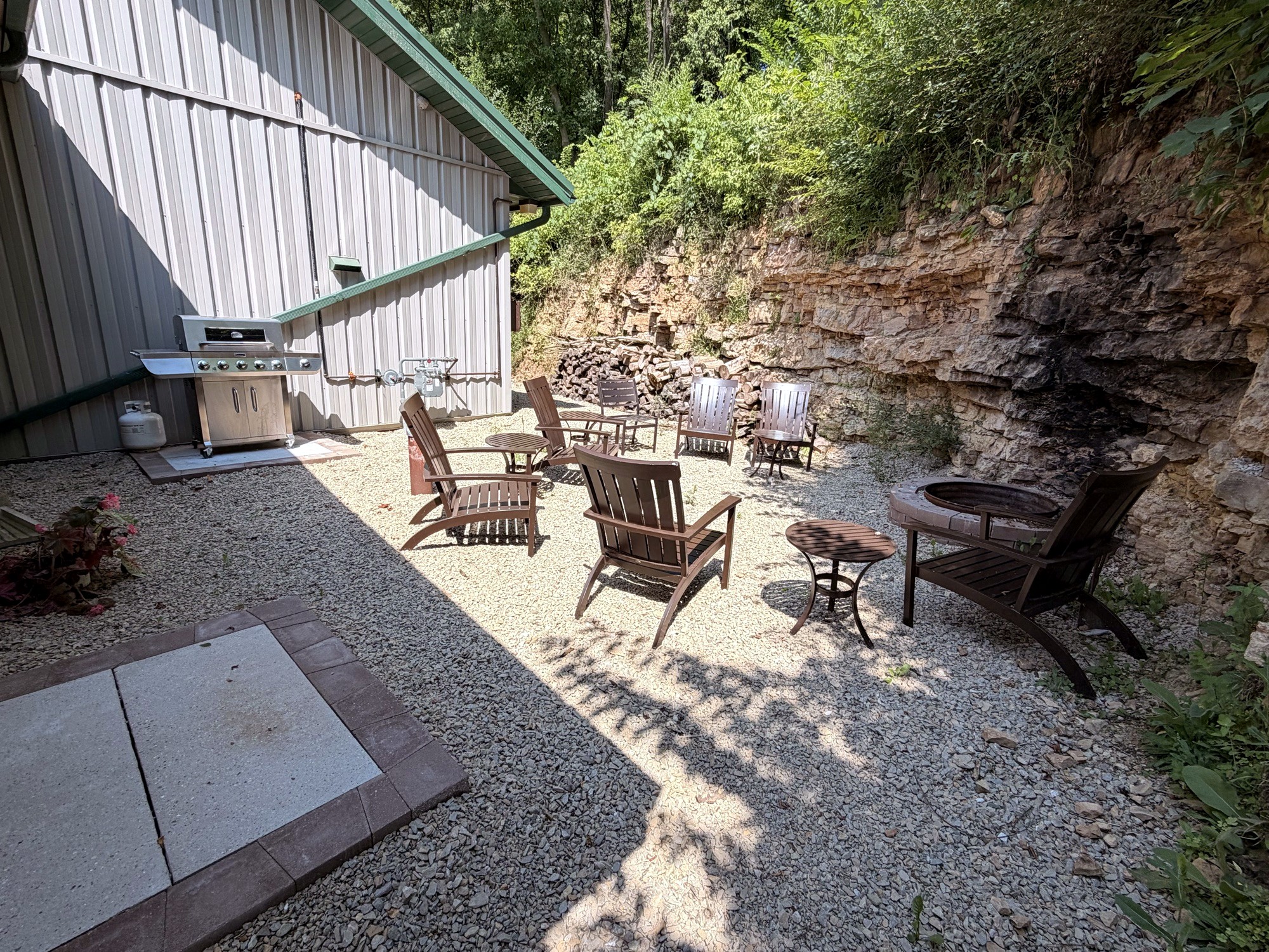The fire pit under the limestone bluff: wooden chairs on the gravel, the grill by the building, trees above the rock wall